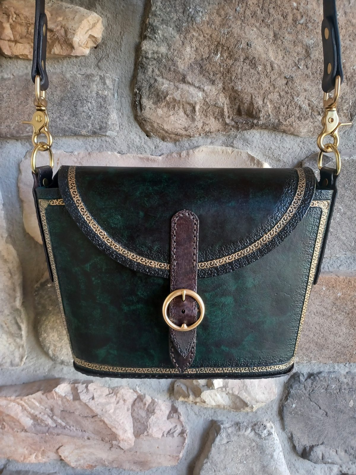 Emerald hand-tooled leather satchel with brass buckle, hung against a stone wall